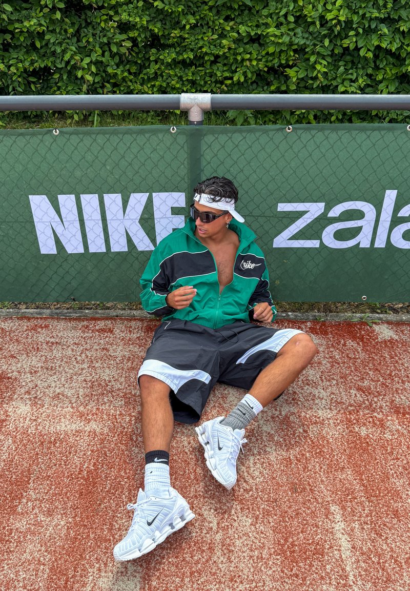 Jeune homme en veste verte Nike, short noir et baskets blanches, assis sur une piste rouge, portant des lunettes de soleil et un bandeau, près d'un panneau Nike.
