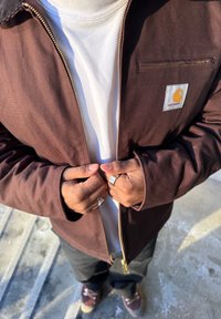 Άτομο που φοράει καφέ μπουφάν Carhartt πάνω από λευκό πουκάμισο, κλείνοντας το φερμουάρ, στέκεται σε εξωτερικά σκάλες από σκυρόδεμα κάτω από τον ήλιο.