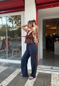 Ung kvinna med uppsatt hår, iklädd en brun ärmlös topp, mörkblå vida byxor, svarta sandaler och bär en mönstrad handväska, stående utanför ett café.