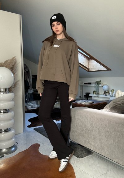 Sudadera marrón con logo, pantalones ajustados negros y zapatillas blancas con rayas negras. El fondo presenta una decoración moderna y un techo inclinado.