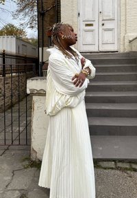 Hvid plisseret kjole med et løst, lagdelt design. Yderligere elementer inkluderer teksturerede fletninger, chunky smykker og en baggrund med trapper og en dør.