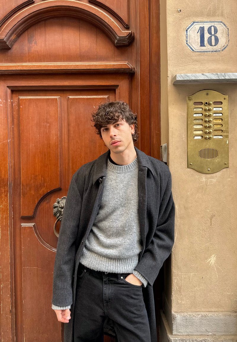 A model leans against a wooden door, wearing a dark coat over a light gray sweater and black jeans. A gold intercom is visible beside him.