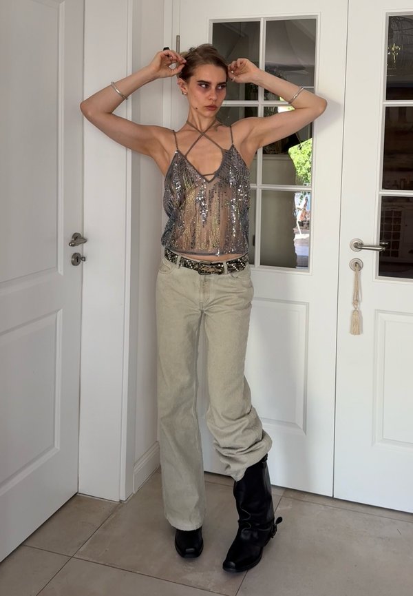 Jeune personne portant un débardeur argenté à sequins, un pantalon clair et une botte noire, les bras levés devant des portes doubles blanches.