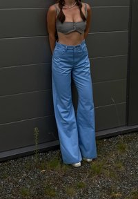 Calças largas de tom azul claro com uma textura suave, combinadas com um top cinza cropped com botões. A figura está em frente a um fundo cinza.