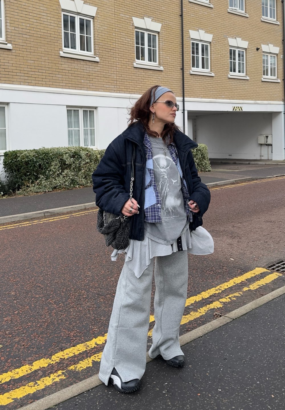 Veste en denim bleu marine surdimensionnée portée sur une tenue superposée grise et noire avec un pantalon de survêtement gris à jambes larges. Accessoires : sac chaîne et lunettes de soleil.