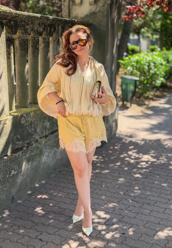 Femeie purtând ochelari de soare, o bluză galbenă largă, pantaloni scurți cu dantelă și pantofi cu toc ascuțit, stă pe un trotuar însorit ținând un clutch.