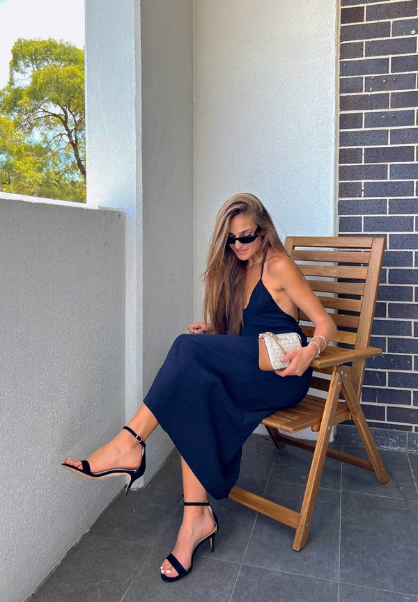 Mujer con cabello largo, vestida con un vestido negro, tacones negros y gafas de sol, sentada en una silla de madera en un balcón, sosteniendo un bolso pequeño.