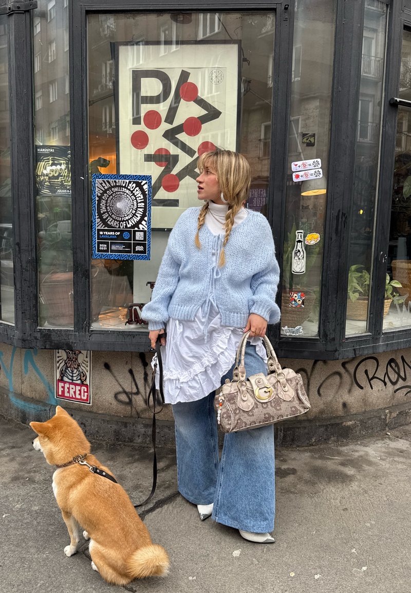 Femme aux cheveux tressés portant un pull bleu et une robe blanche, tenant un sac à main à motifs, debout sur un trottoir avec un chien Shiba Inu en laisse.