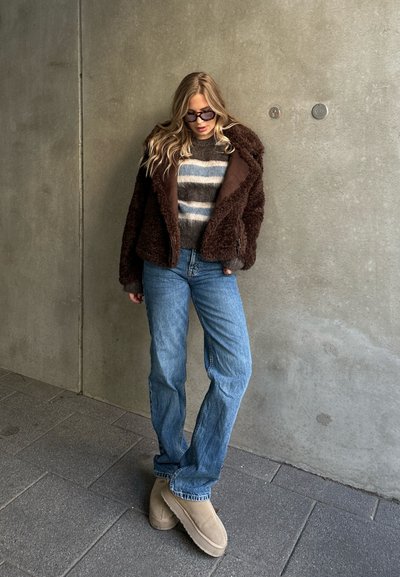 Chaqueta de piel sintética marrón, suéter de rayas gris, jeans azul claros y botines tobilleros color camel. De pie contra una pared de concreto.