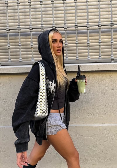 Sudadera negra con cremallera y diseño gráfico, pantalones cortos grises con volantes, bolso plateado con tachuelas y bebida verde. El fondo incluye rejas de metal.