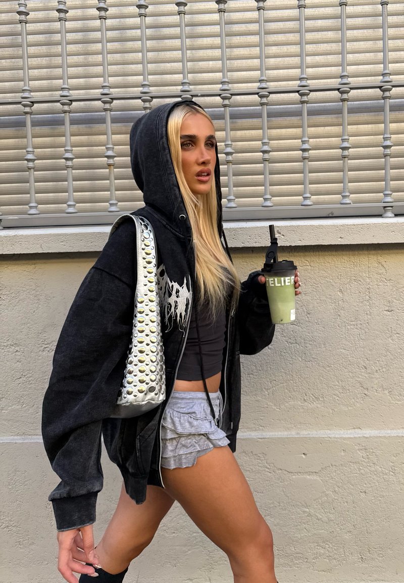 Sudadera negra con cremallera y diseño gráfico, pantalones cortos grises con volantes, bolso plateado con tachuelas y bebida verde. El fondo incluye rejas de metal.