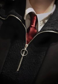 Gros plan d'une personne portant une chemise blanche, une cravate rouge et un pull noir zippé avec un tire-zip circulaire.