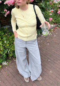 Helles, gelbes, geripptes Cardigan mit Knöpfen, kombiniert mit lockeren grauen Leinenhosen. Die Schuhe haben ein Leopardenmuster. Klare Tasse in der Hand.