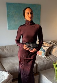 Femme portant une robe en satin violet foncé et un foulard noir, tenant une pochette noire et argentée, debout dans un salon avec un canapé beige.