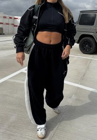 Sort træningsdragt med hvide accenter, kort lynlåstrøje, løse sorte bukser med sideløbende stribe og hvide sneakers med grå detaljer.