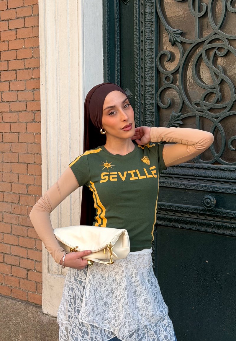 Femme portant un hijab brun foncé, un t-shirt vert "Seville", des manches beiges transparentes, une jupe en dentelle blanche, tenant un sac à main blanc, debout près d'une porte noire ornée.