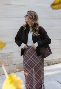 Cardigan castanho, texturizado, sobre uma blusa branca, combinado com uma saia xadrez roxa apresentando uma fenda lateral. Uma bolsa escura com um detalhe em corrente.