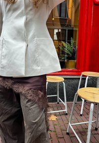 Veste blanche cintrée avec fermeture éclair, associée à un pantalon marron orné de dentelle. En arrière-plan, un cadre de fenêtre rouge et des tabourets en bois.