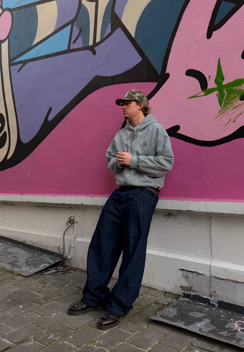Jeune homme portant un sweat à capuche gris, une casquette camouflage et un jean large foncé, appuyé contre un mur orné d'un graffiti abstrait coloré.