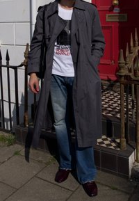 Grå trenchcoat, hvit grafisk t-skjorte, lyseblå flarejeans og mørk burgunder sko med krokodillemønster; stående på mønstrete asfalt.