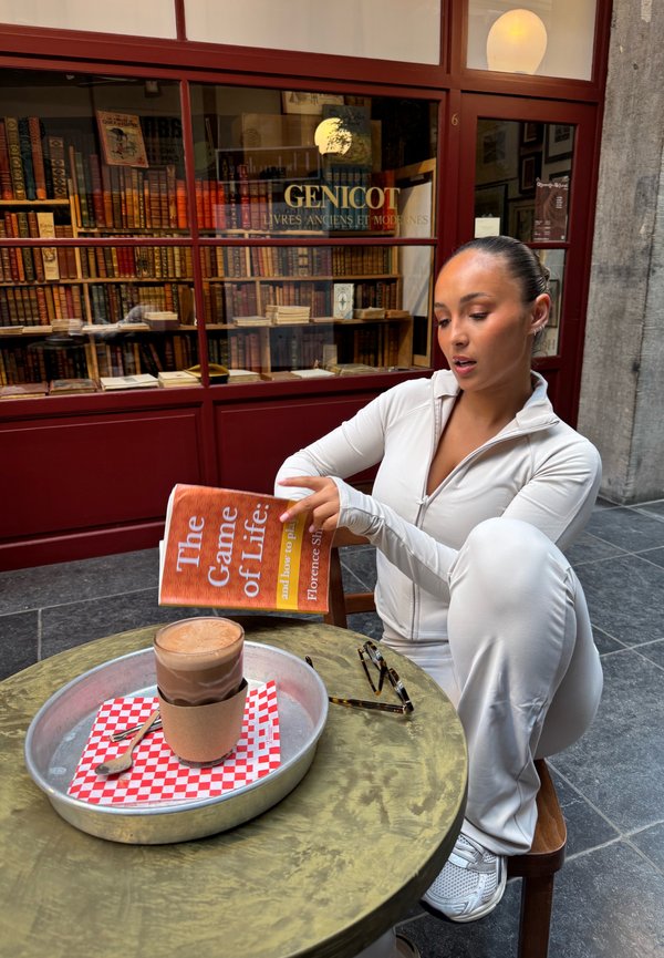 Book titled "The Game of Life" held by a person in a white zip-up jacket, seated at a round outdoor table with a metal tray and a dessert.
