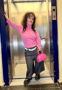 Rozi džemper s rolkom, sive široke hlače, crne platforme. Drži malu rozu torbicu; stoji u liftu.