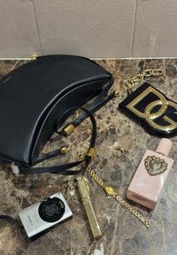 Μαύρη τσάντα με σχήμα σεληνοειδούς, μαύρο μπρελόκ με επιχρυσωμένες λεπτομέρειες, ροζ μπουκάλι αρώματος, χρυσό ρολόι, δαχτυλίδι, χρυστό κραγιόν και ασημένια ψηφιακή κάμερα πάνω σε μαρμάρινη επιφάνεια.