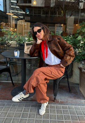 Mujer con gafas de sol, chaqueta marrón con manchas y pantalones cargo color óxido sentada en una mesa de una cafetería al aire libre con una taza de café y un bolso beige.