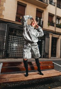 Tenue métallique argentée composée d'une veste ample et d'un short. Le mannequin porte des bottes noires à talons aiguilles montant jusqu'aux cuisses et adopte une pose dynamique sur un banc en bois.