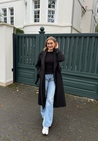 Black long coat over a black top, paired with light blue flared jeans and white trainers, standing in front of a green gate.