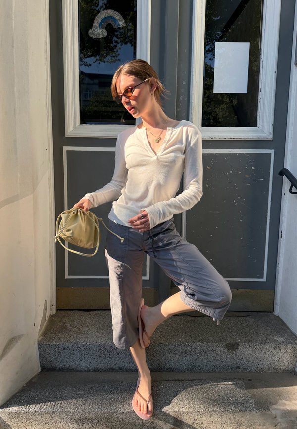 Junge Frau mit Sonnenbrille, weißem Langarmshirt und grauer, verkürzter Hose steht auf einem Fuß und hält eine gelbe Tasche vor einer Tür.