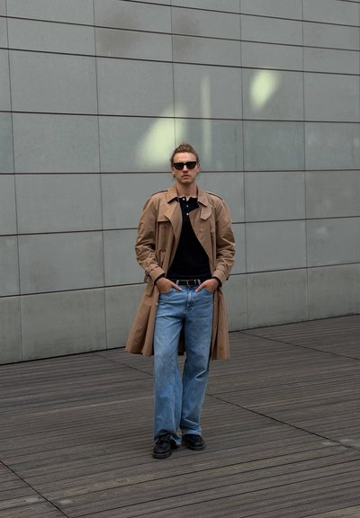 Hombre con gafas de sol, gabardina beige, camisa negra, vaqueros azules y zapatos negros, de pie con las manos en los bolsillos sobre suelo de madera cerca de una pared gris.