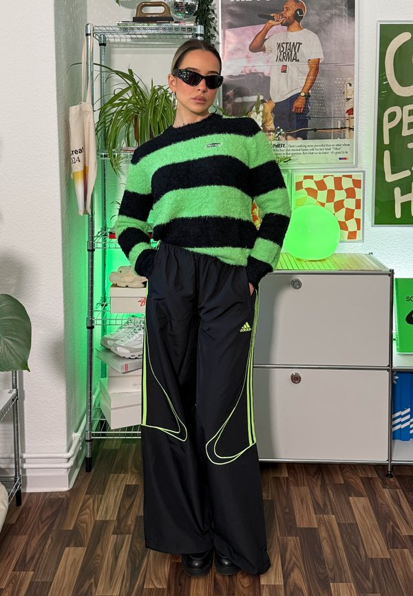 Un pull flou rayé vert et noir à manches longues, associé à un pantalon de survêtement noir avec des accents néon et un logo Adidas.