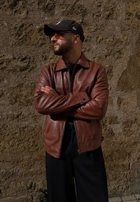 Chaqueta de cuero marrón con cierre frontal, cuello apuntado y acentos contrastantes en negro. Modelo lleva una gorra negra y gafas, de pie frente a una pared texturizada.