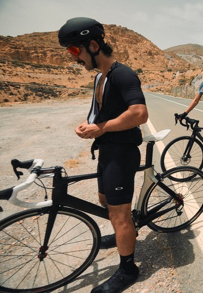 Un ciclista en un traje aerodinámico negro con cremallera frontal, que lleva un casco negro y gafas de sol con tinte naranja, se encuentra al lado de una bicicleta de carretera negra.
