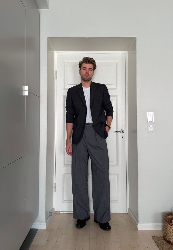 Homme portant un blazer noir, une chemise blanche, un pantalon gris à jambes larges et des chaussures noires, debout à l'intérieur devant une porte blanche, la main dans la poche.