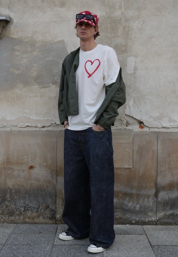 Joven con camiseta blanca con un corazón rojo, chaqueta verde colgada de los hombros, vaqueros anchos azules, gorra roja con gafas de sol, de pie contra una pared.