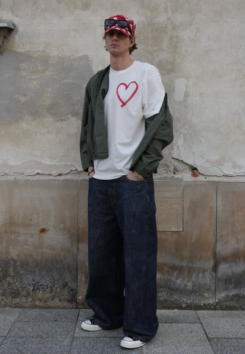 Jeune homme portant un t-shirt blanc avec un cœur rouge, une veste verte tombant des épaules, un jean large bleu, une casquette rouge avec des lunettes de soleil, debout contre un mur.