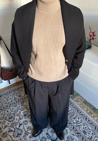 Sort frakke over en beige rullekravesweater, kombineret med løstsiddende sorte bukser og skinnende sorte sko, der viser et lagdelt mod look.