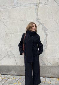 Vrouw in zwarte jas, sjaal en wijde broek staande tegen een gebarsten beige stenen muur met vallende gele bladeren op het trottoir.