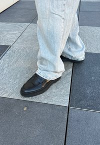 Μαύρες δερμάτινες πατούσες με λεία επιφάνεια και λουρί στη πάνω πλευρά, σε συνδυασμό με ανοιχτό μπλε, ελαφρώς φθαρμένα τζιν παντελόνια.