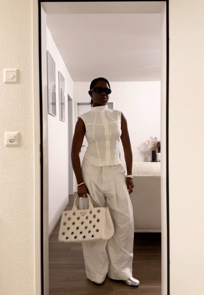 Mujer con top blanco sin mangas y pantalones anchos sostiene un bolso blanco con recortes circulares, lleva gafas de sol oscuras en interiores.
