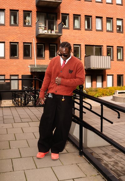 Cardigan de punto rojo con un logo, camisa blanca y corbata roja. Pantalones de pierna ancha negros y zapatos rojos brillantes. Entorno urbano con edificios de ladrillo.