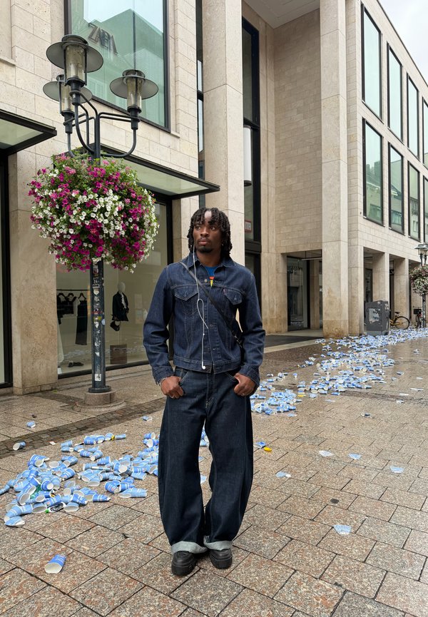 Uomo con giacca e pantaloni di jeans in piedi per strada con bicchieri di carta sparsi vicino a un edificio con grandi finestre e cesti di fiori appesi.
