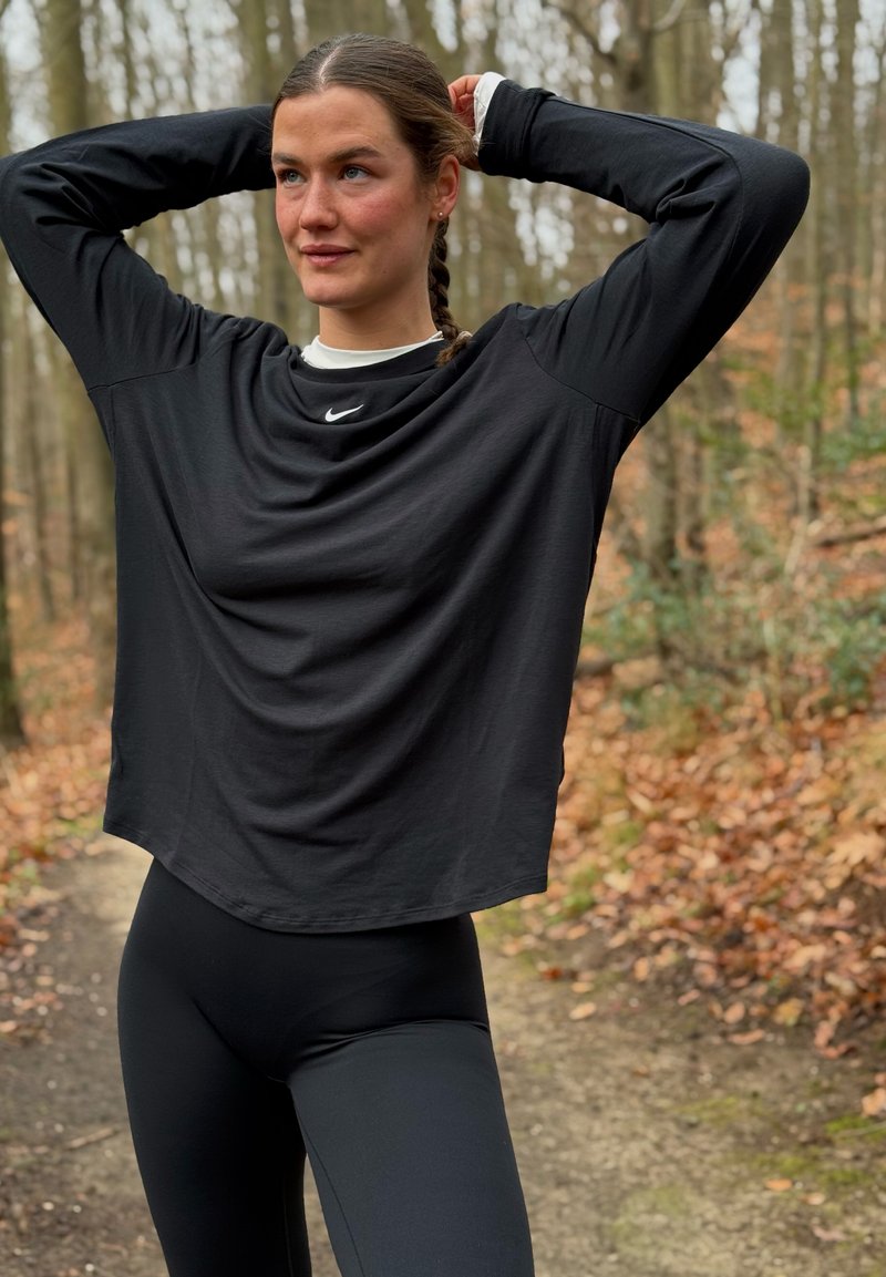T-shirt de sport noir à manches longues avec ourlet incurvé et logo Nike, assorti à un legging noir, dans un cadre extérieur forestier avec des feuilles d'automne.