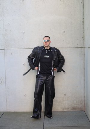 Chaqueta de cuero negra con herrajes plateados, camiseta atlética negra con acentos blancos y pantalones anchos de imitación cuero negros. Gafas de sol espejadas plateadas.