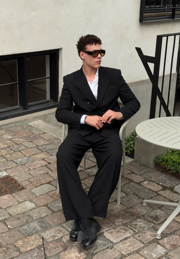 Mann in schwarzem Anzug und Sonnenbrille sitzt im Freien auf einem Stuhl neben einem runden Metalltisch auf einem gepflasterten Hof.