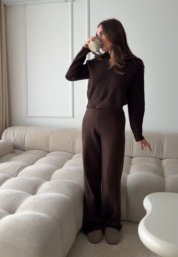 Vrouw in bruine gebreide kleding staat bij een beige gestoffeerde bank, drinkt uit een keramische kom in een minimalistische, neutraal getinte woonkamer.