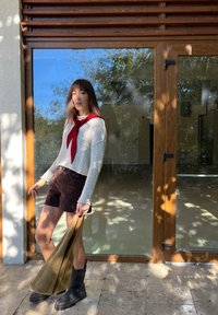 Gebreide crèmekleurige trui met een rode sjaal, bruine suède shorts en zwarte enkellaarzen. Houdt een groenachtige-bruine tas vast. Achtergrond met glazen deur en natuurlijk licht.