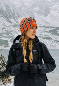 Crna vodootporna jakna s patentnim zatvaračem sprijeda, uparen s prugastom narančastom i crnom pletenom kapom. Nosim crne rukavice, kosa je u pletenicama.